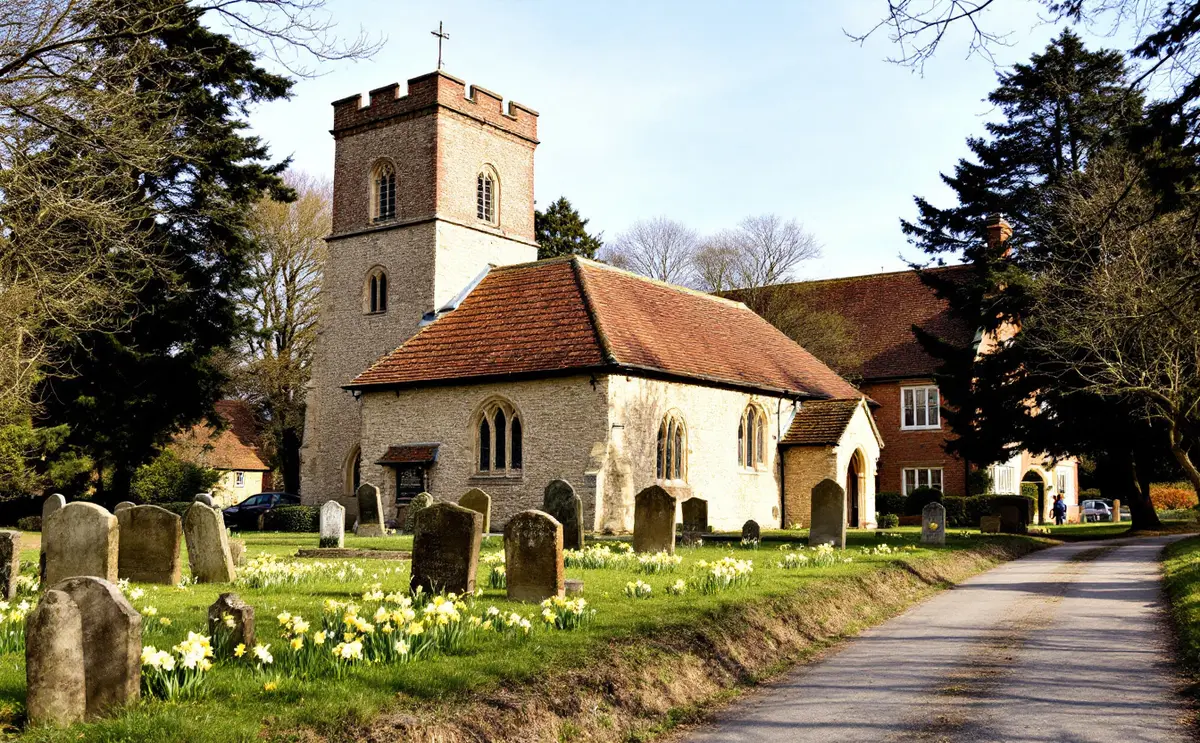 A flint church with a short brick tower and small spire beside trees and a brick house, early spring.